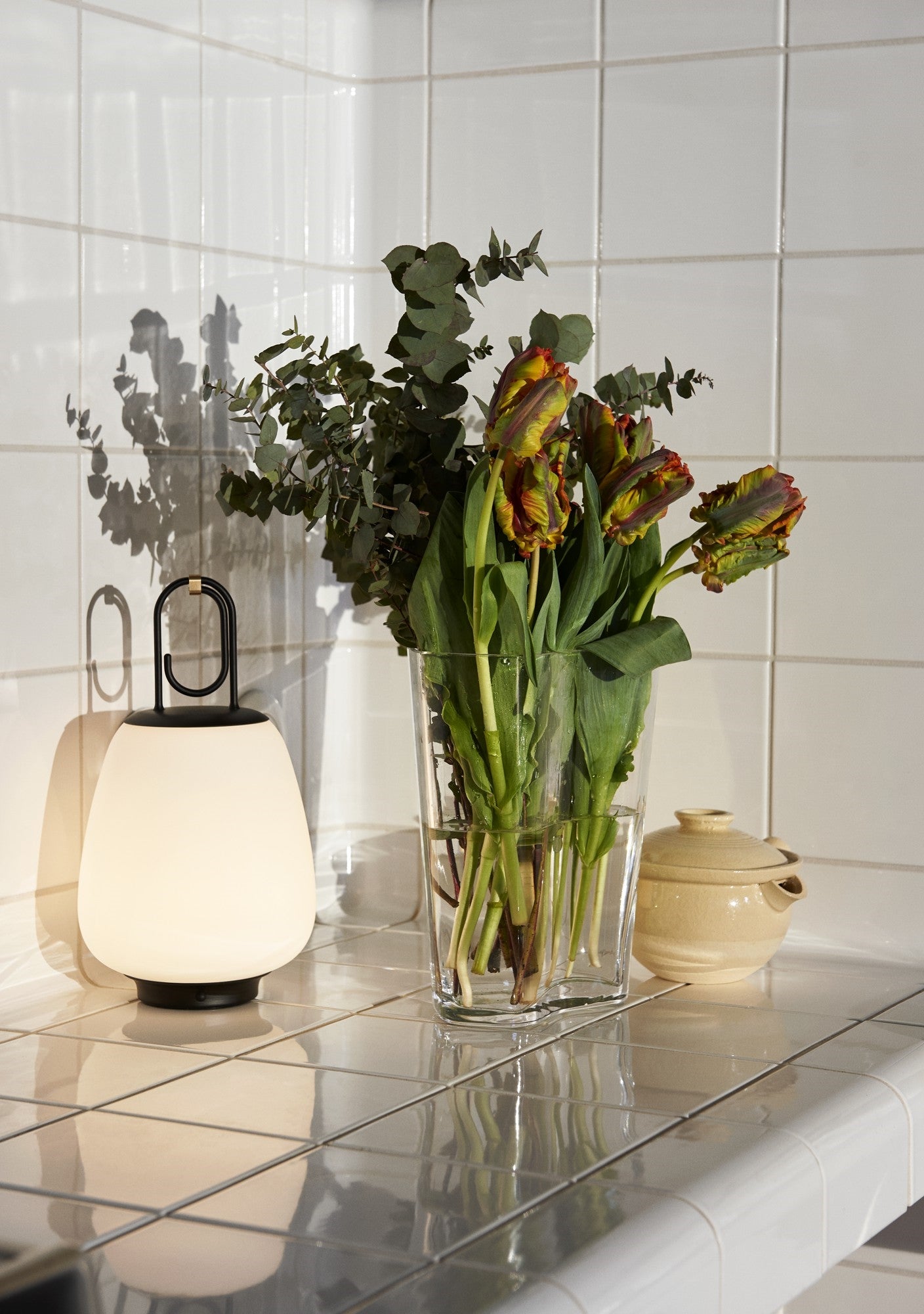 Vase mit Tulpen und Eukalyptus auf weißem Fliesentisch, neben moderner Tischlampe.