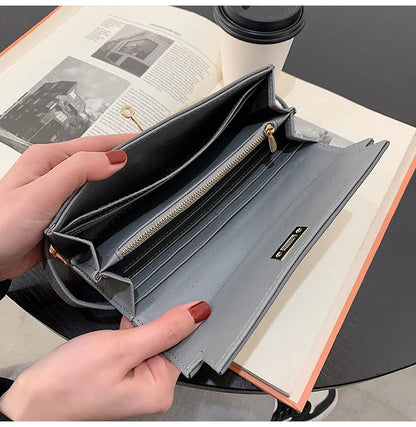 Graue Damen-Geldbörse aus Leder mit Reißverschluss, geöffnet auf Tisch mit Buch und Kaffeetasse.