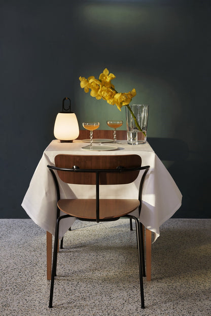Moderner Esstisch mit weißer Tischdecke, gelben Blumen, Glasvase, Lampe und Holzstuhl.