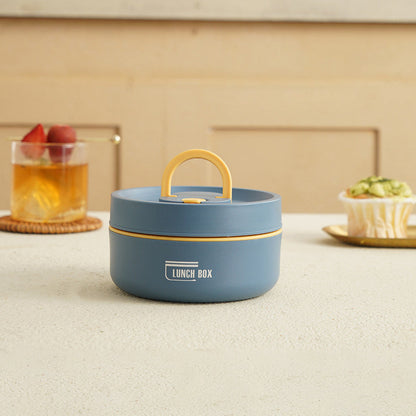 Blaue runde Lunchbox mit gelbem Griff auf Tisch, neben Getränk und Muffin.