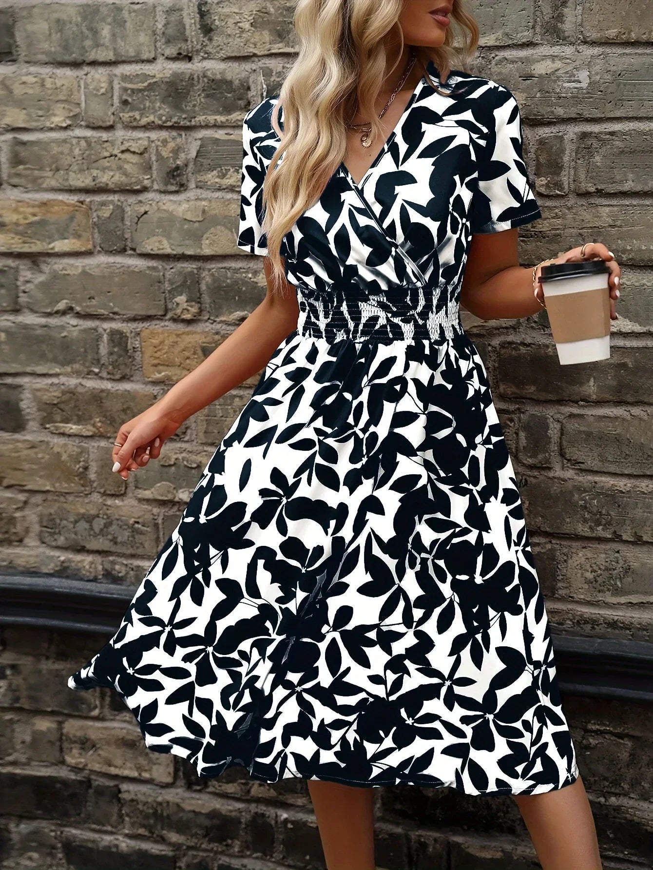 Frau im schwarz-weiß geblümten Sommerkleid mit Kaffeebecher vor Ziegelwand.