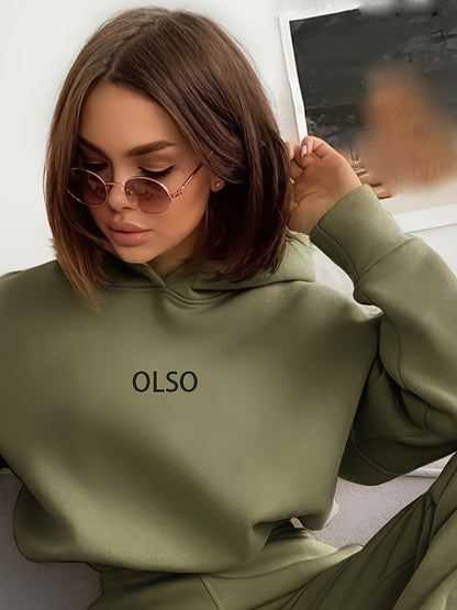 Frau in olivgrünem Hoodie mit "OLSO"-Aufdruck, trägt Sonnenbrille, sitzt auf Sofa.