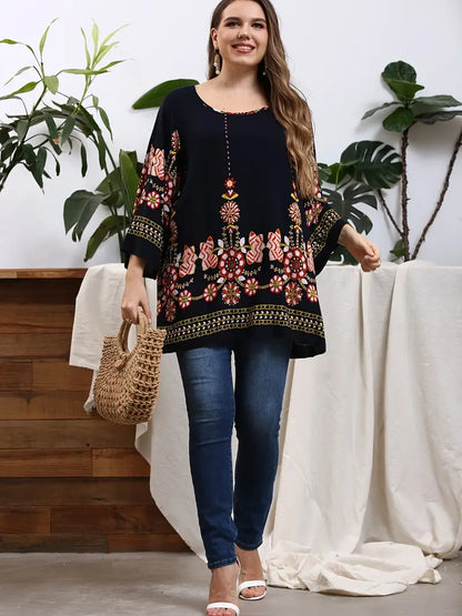 Frau in schwarzer Tunika mit Blumenmuster, Jeans und Strohtasche, lächelnd, vor Pflanzen.