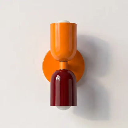 Vägglampa med orange övre skärm och orange väggplatta samt blank vinröd nedre skärm, uppsatt på en ljus vägg. 25