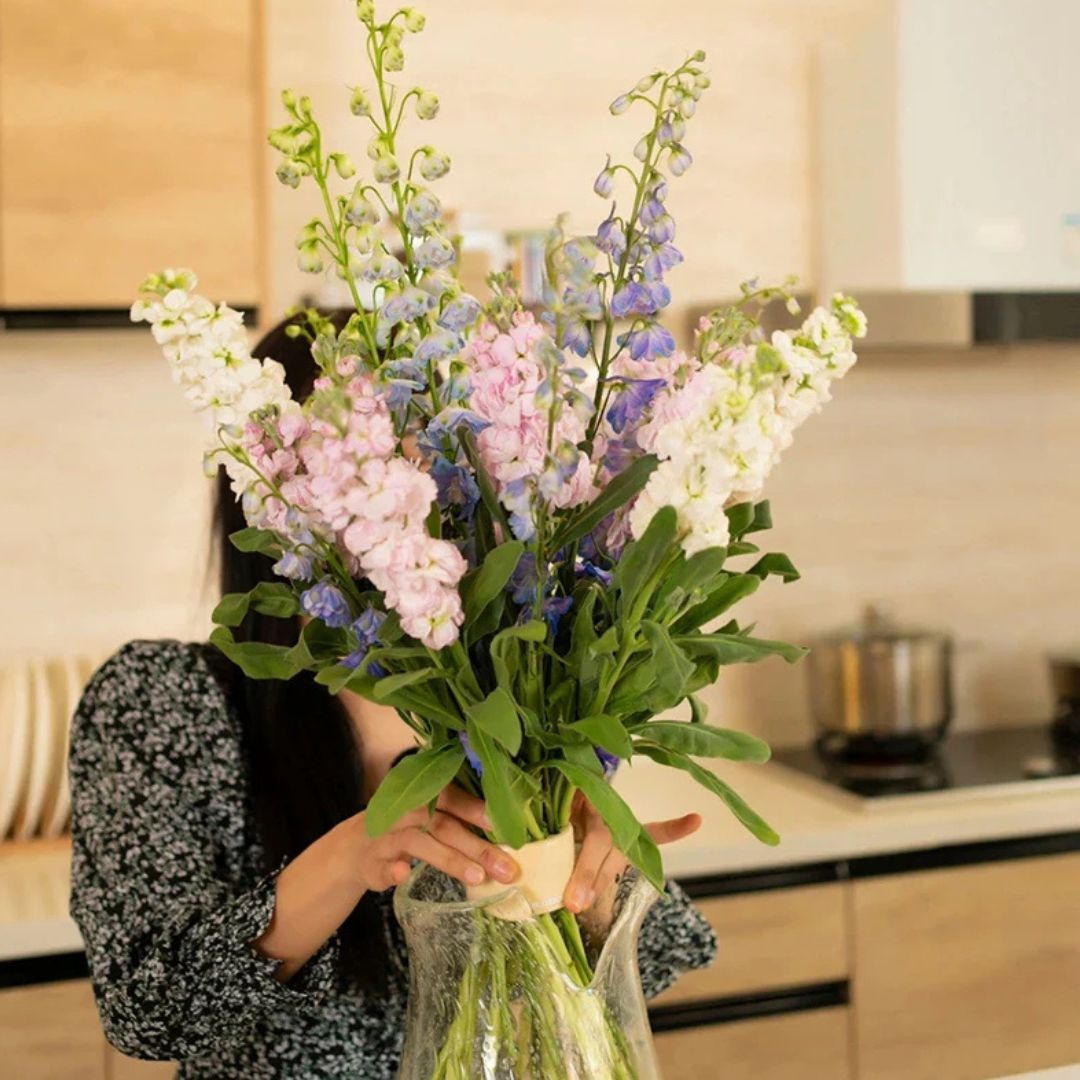 Person i mönstrad tröja håller en hög, upprättstående bukett med rosa, vita och blå blommor i en glasvas i köket. 2
