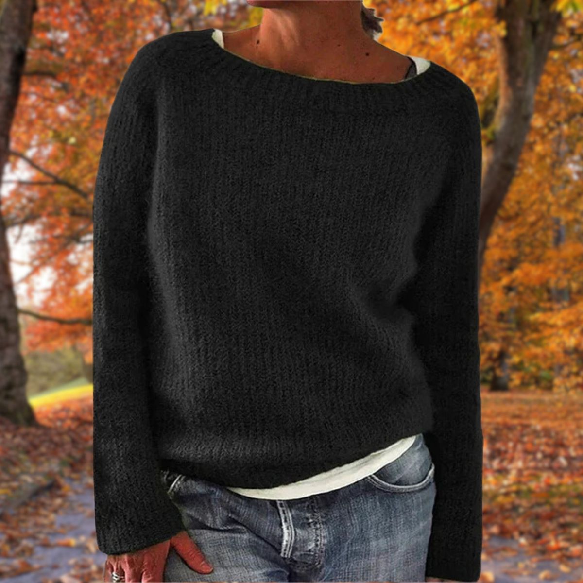 Svart stickad tröja med rund hals, bärd av en modell över vitt linne och jeans med lövfärgad höstpark i bakgrunden. 2