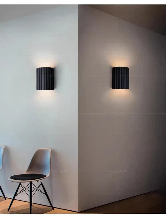 Schwarze Wandleuchte aus Metall, modernes Design, beleuchtet, neben grauem Stuhl, Innenraum.