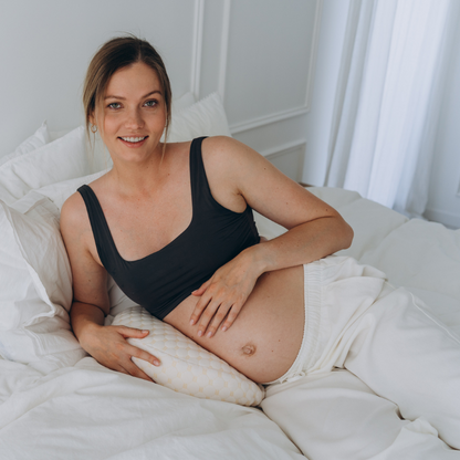 Schwangere Frau mit Schwangerschaftskissen im Bett, lächelnd, in schwarzem Top und weißer Hose.