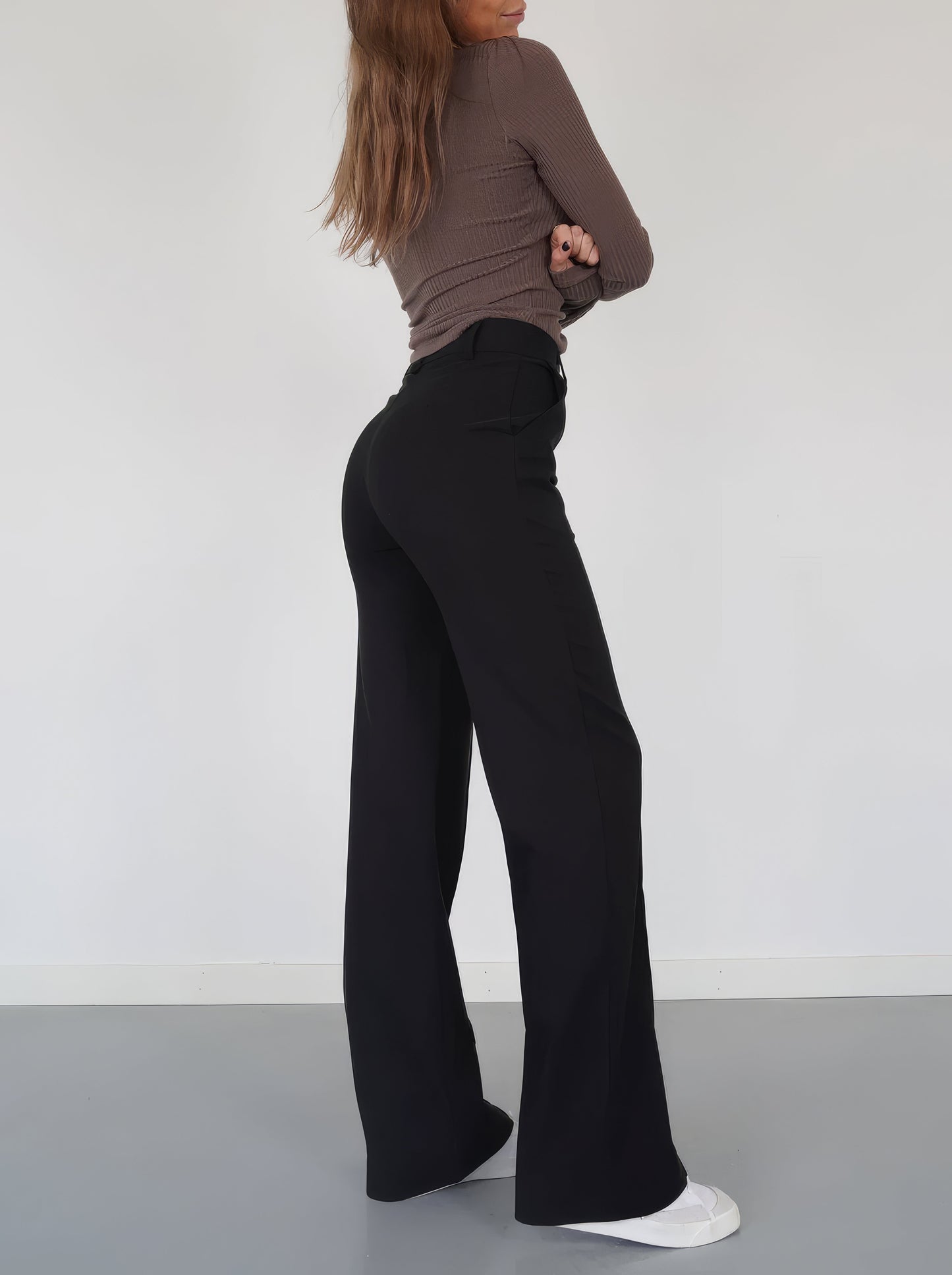 Frau in schwarzer Schlaghose und braunem Oberteil, seitlich, weiße Sneaker, minimalistischer Stil.