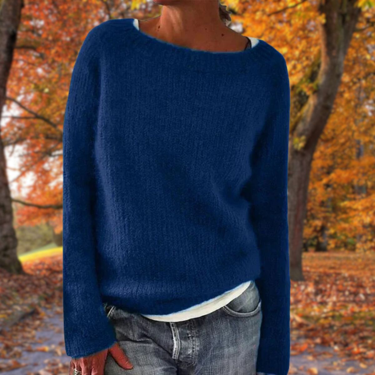 Marinblå stickad tröja med rund hals, bärd av en modell över vitt linne och jeans bland gulorange höstträd. 3