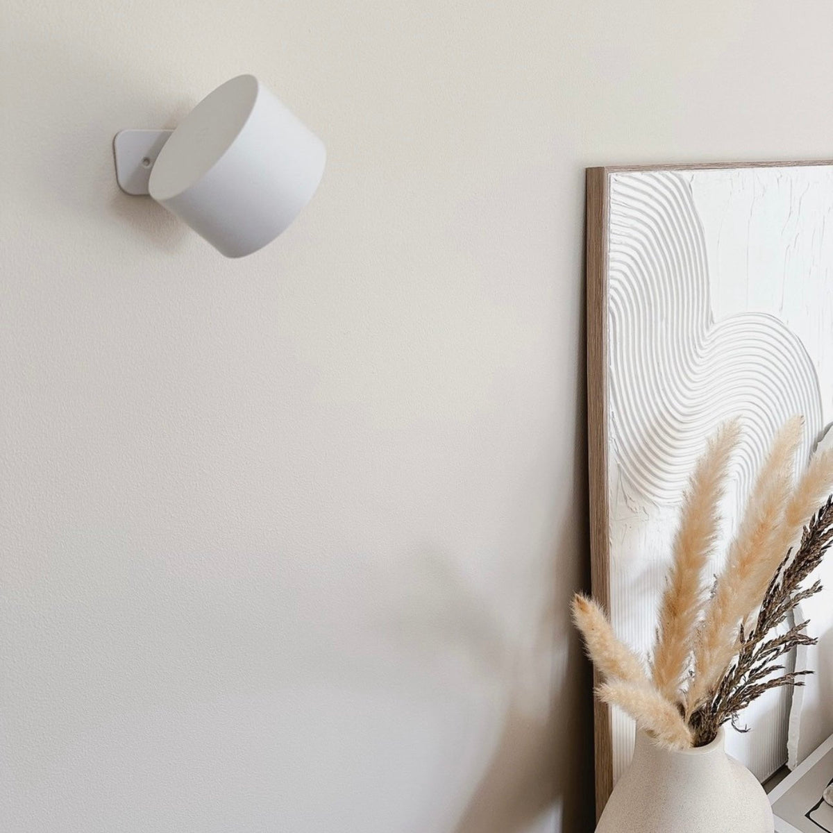 Släckt vit vägglampa monterad på en ljus vägg, med en inramad strukturerad tavla och torkade pampasgräs i en vas bredvid. 17