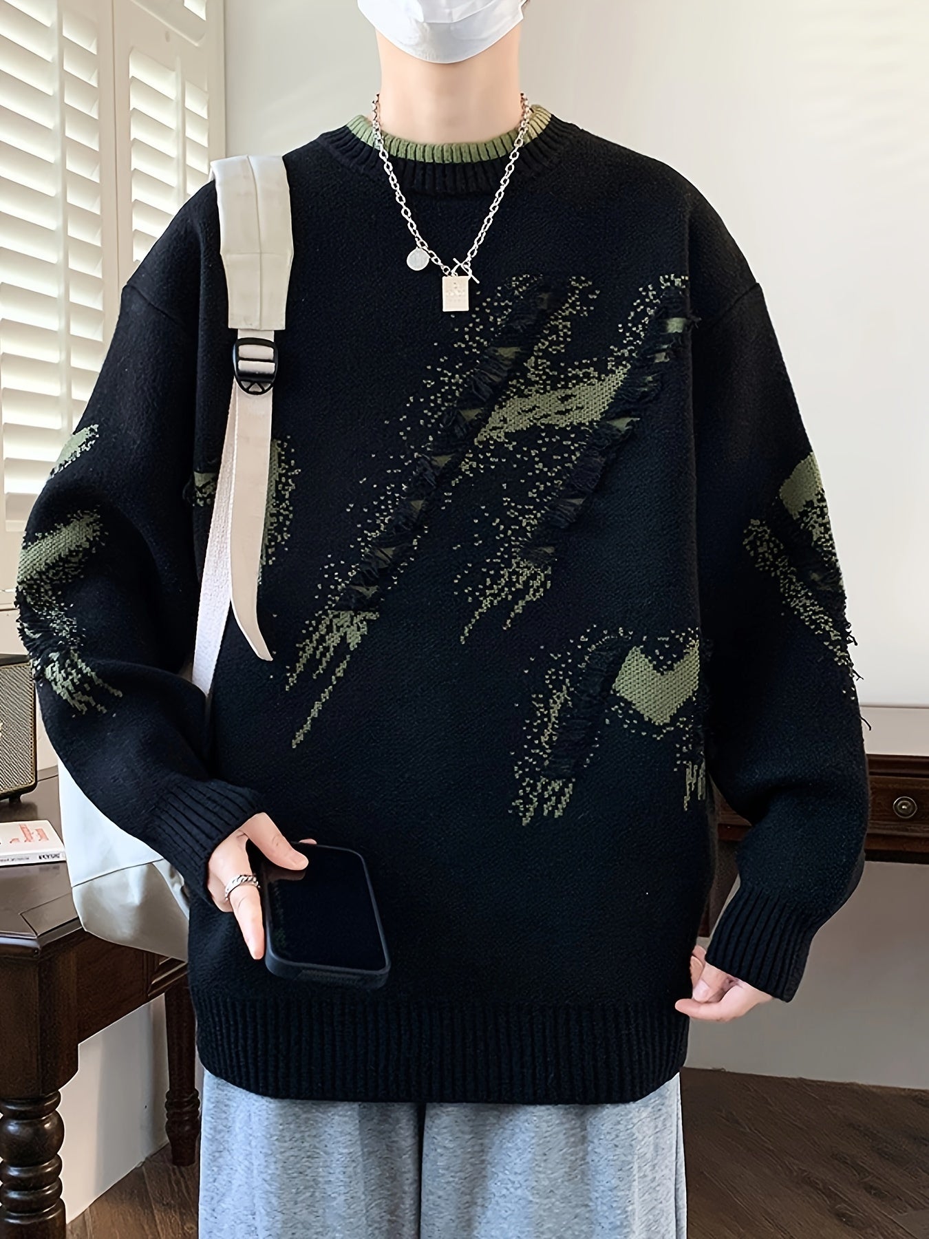 Mann in schwarzem Strickpullover mit grünem Muster, hält Smartphone, trägt weiße Maske.