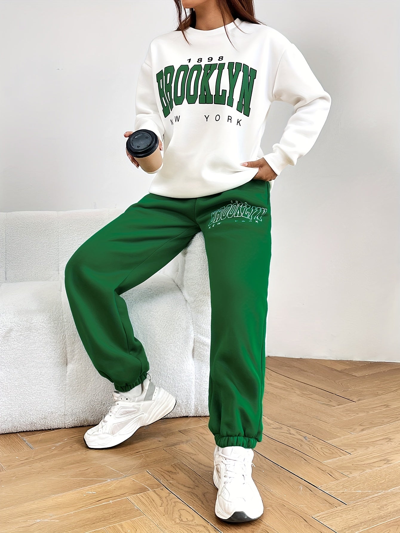 Frau in weißem Brooklyn-Pullover und grüner Jogginghose mit Kaffeetasse, lässige Mode.