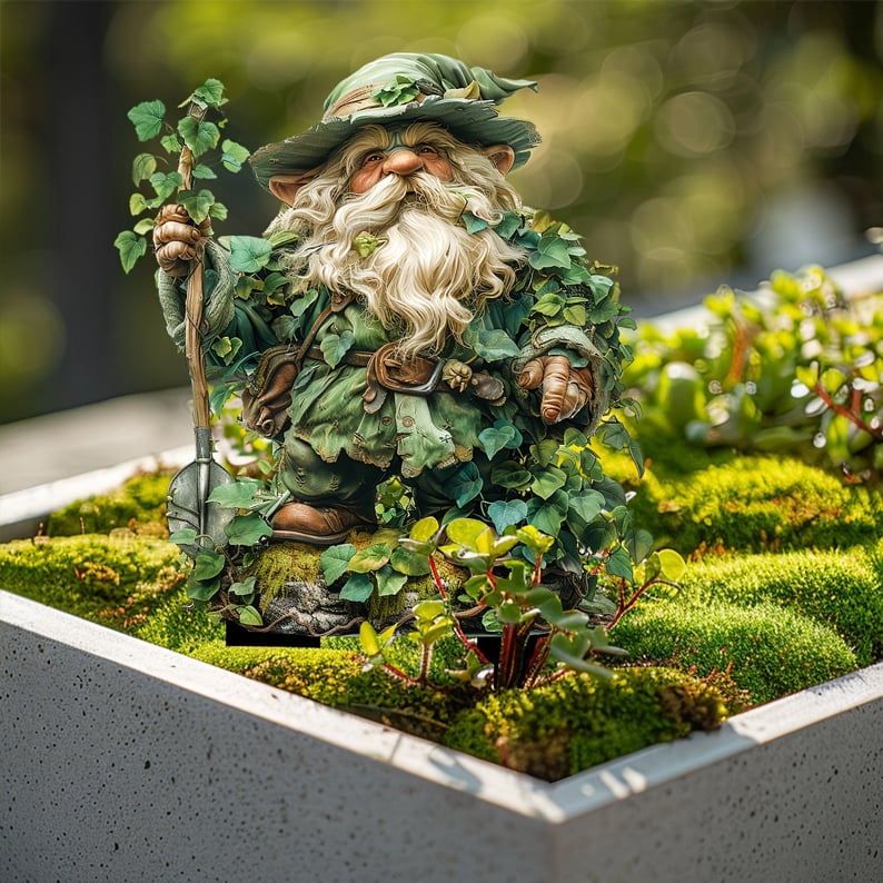 Bladklädd tomte med spade, stående i en grå planteringslåda full av mossa och små gröna växter. 15