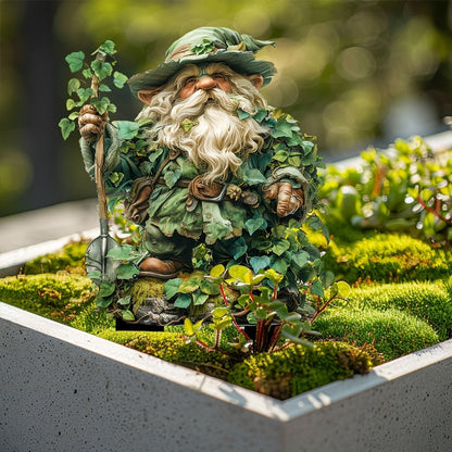 Bladklädd tomte med spade, stående i en grå planteringslåda full av mossa och små gröna växter. 15