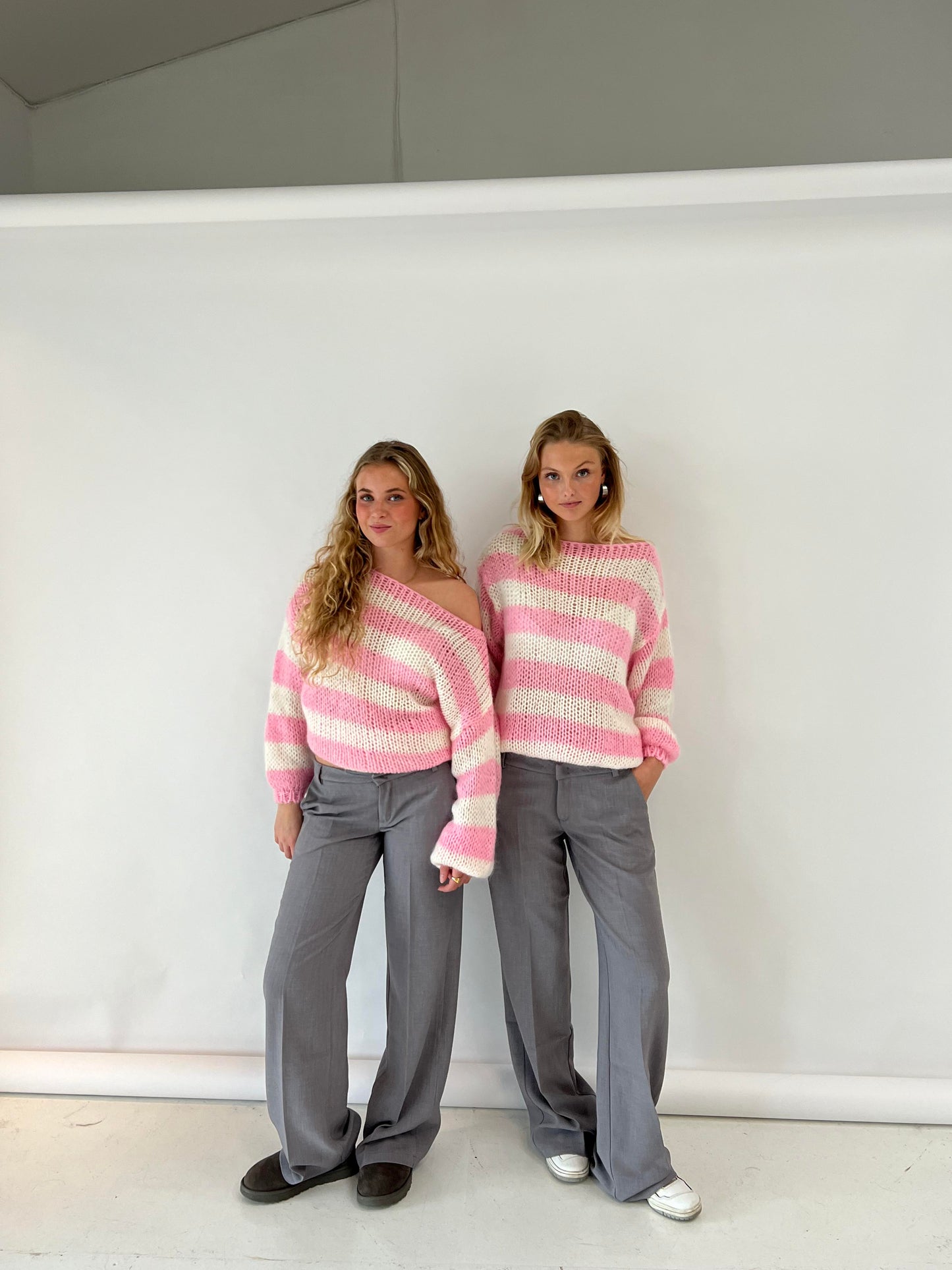 Zwei Frauen in gestreiften rosa Pullovern und grauen Hosen vor weißem Hintergrund.