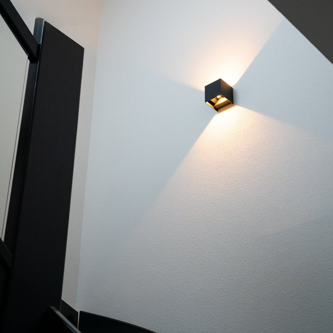 Svart kubformad vägglampa i ett trapphus, sedd underifrån, med varmt ljus som sprids över en vit väg. 2