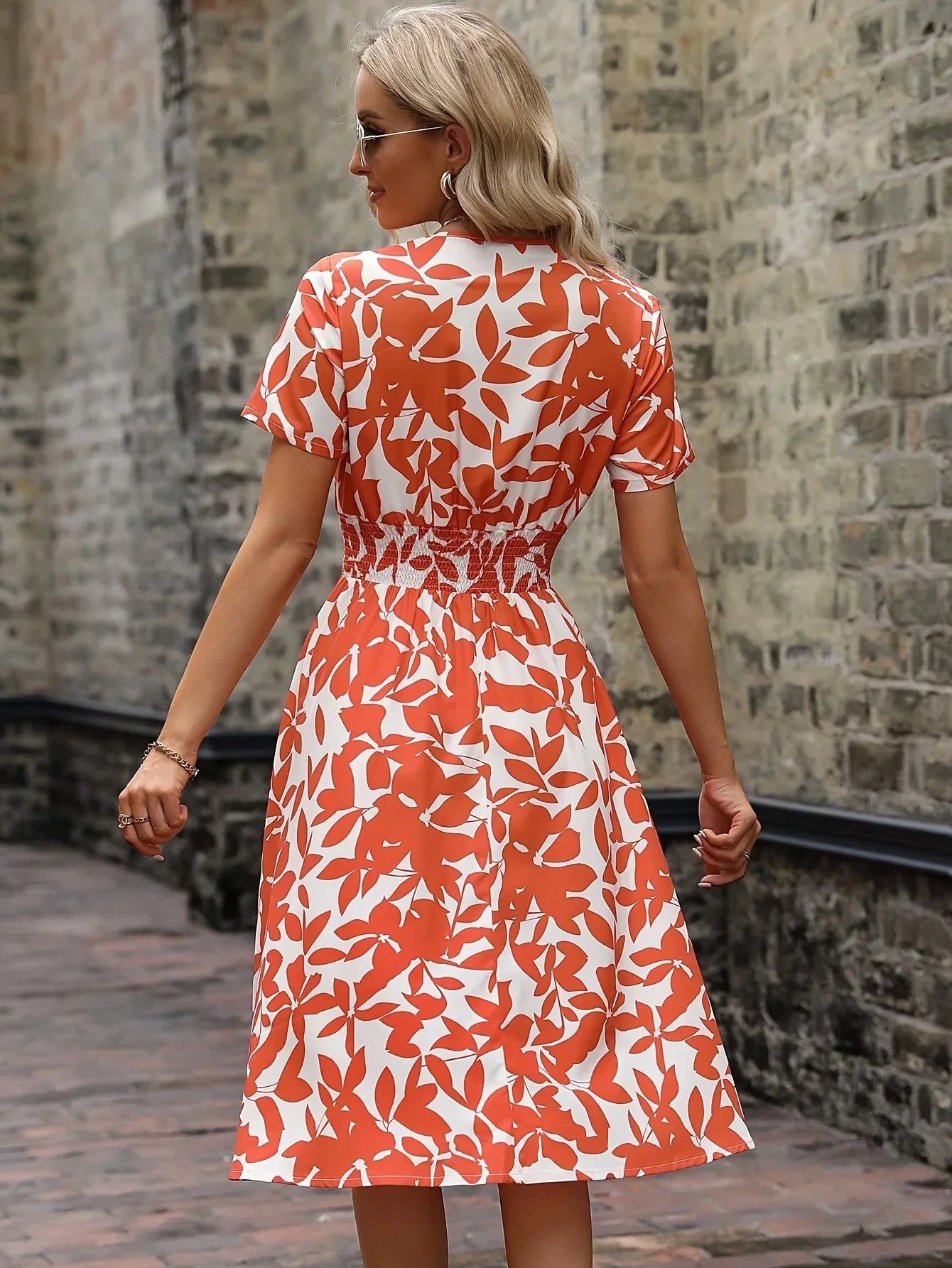 Frau in orange-weißem Sommerkleid mit Blattmuster, steht vor einer Steinmauer.