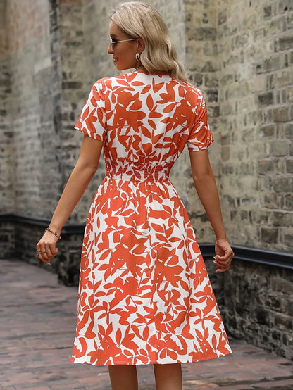 Frau in orange-weißem Sommerkleid mit Blattmuster, steht vor einer Steinmauer.