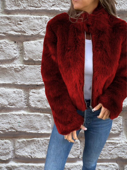 Frau in roter Kunstpelzjacke, weiße Bluse, blaue Jeans, vor Ziegelwand.