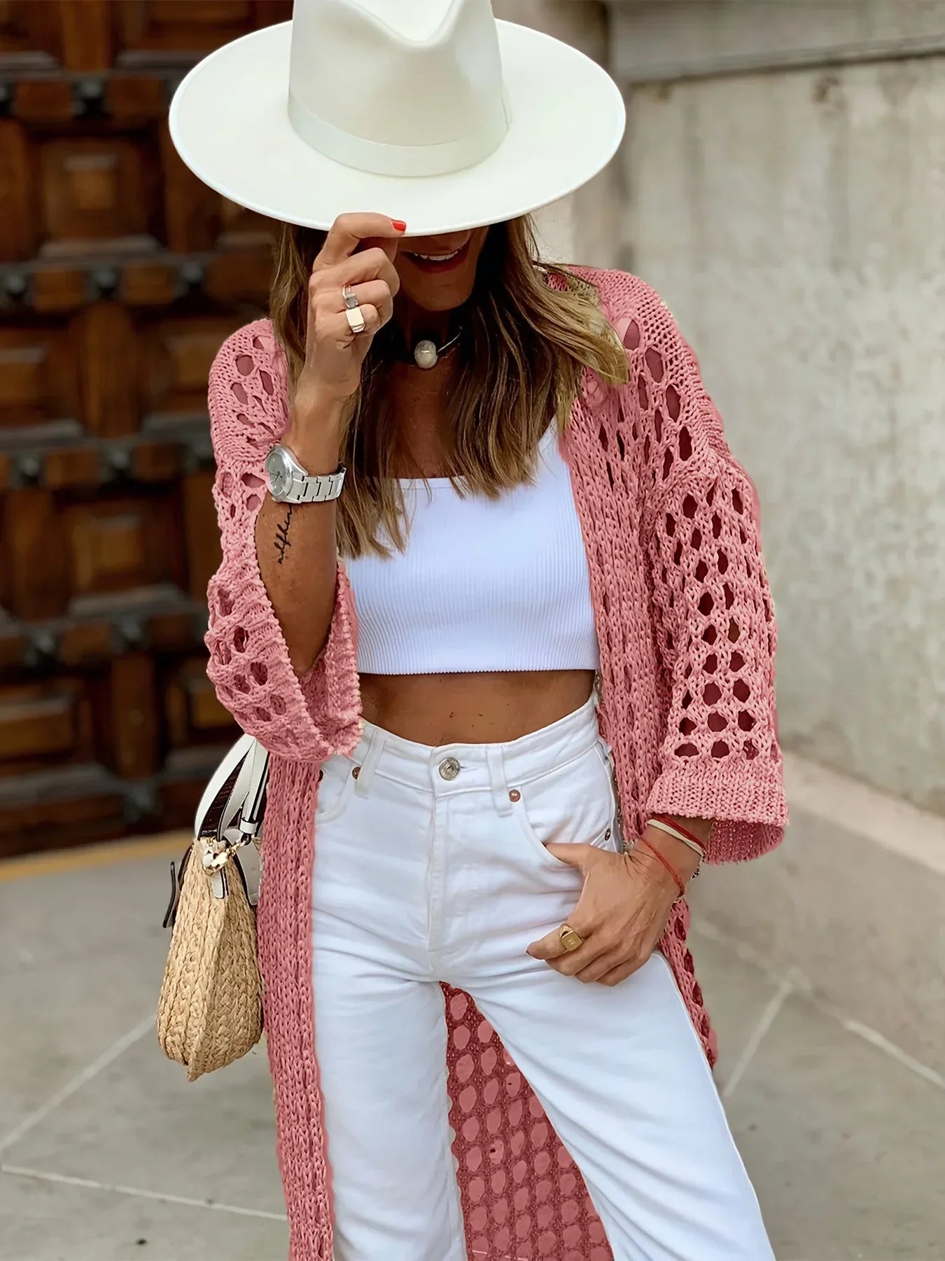 Frau in rosa Strickjacke, weißem Hut und weißer Hose, hält Strohtasche, modisch, urban.