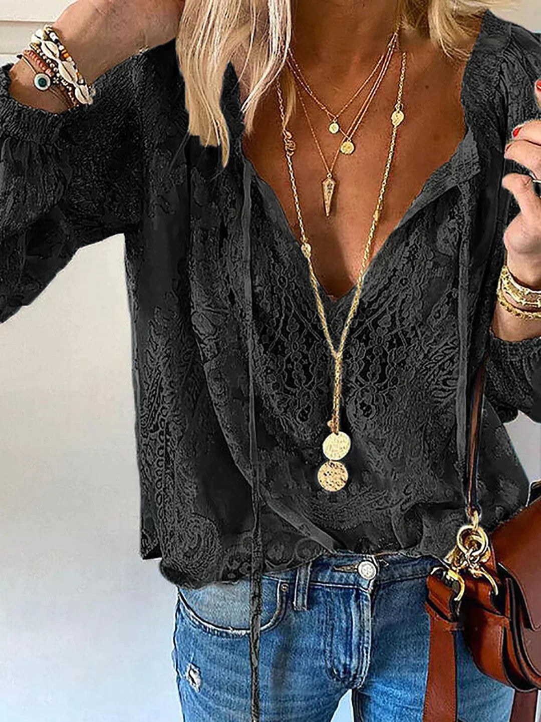 Frau in schwarzer Spitzenbluse, blaue Jeans, goldene Halsketten, braune Ledertasche.