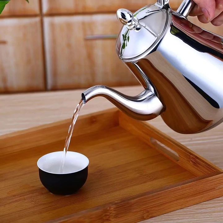 Silberne Edelstahl-Teekanne gießt Tee in schwarze Tasse auf Bambus-Tablett.