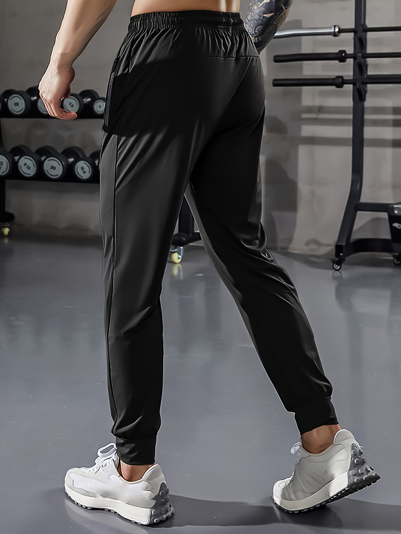 Mann in schwarzer Sporthose im Fitnessstudio, Hanteln im Hintergrund, weiße Turnschuhe.
