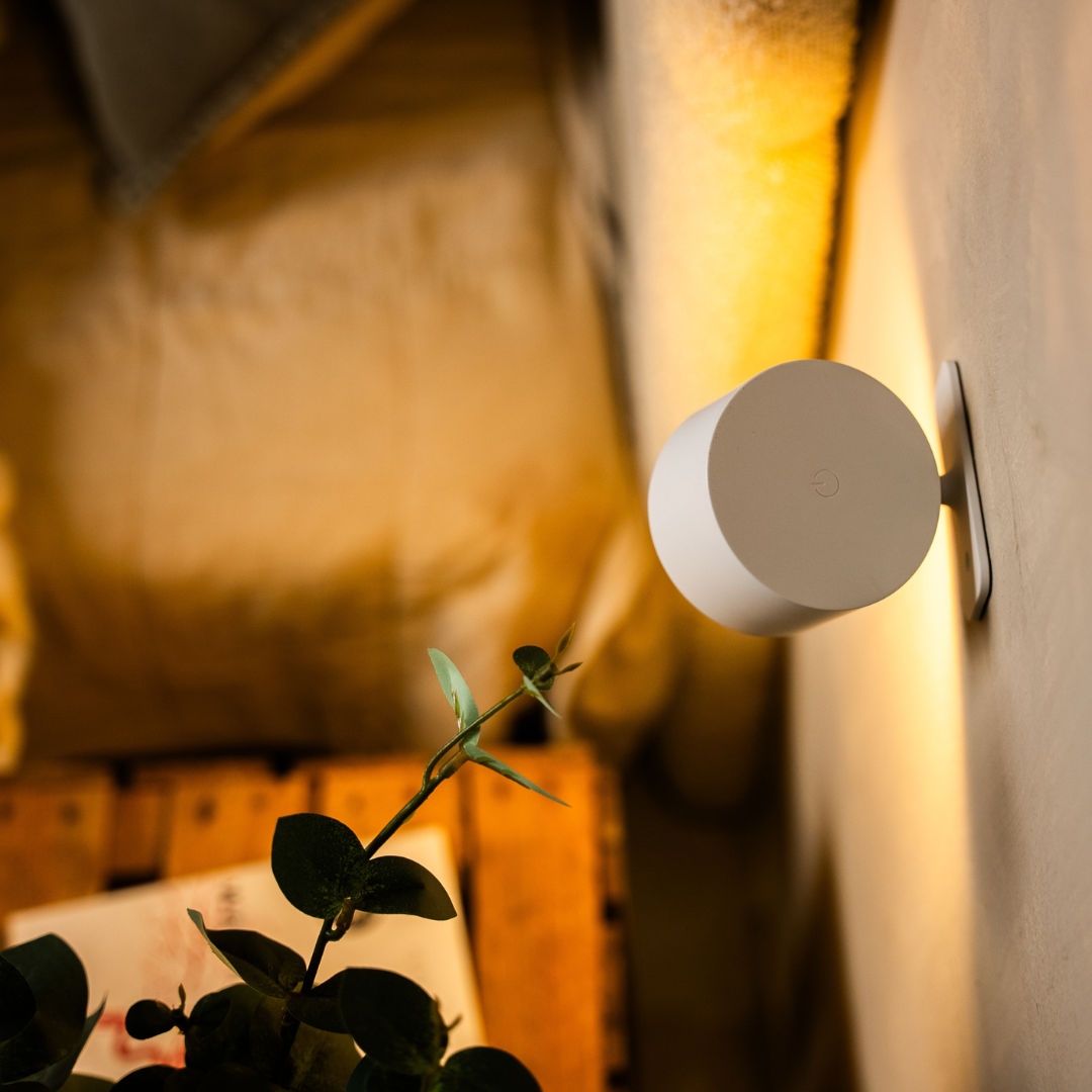 Baksidan av en vit vägglampa i närbild, med en liten strömknapp på den runda ytan och väggfästet synligt. 13