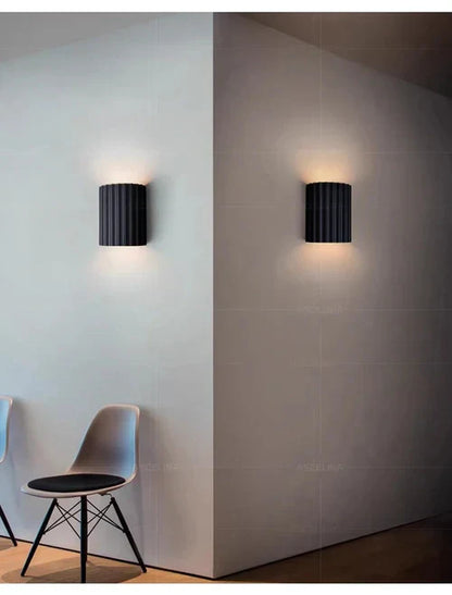 Schwarze Wandleuchte aus Metall, modernes Design, beleuchtet, neben grauem Stuhl.