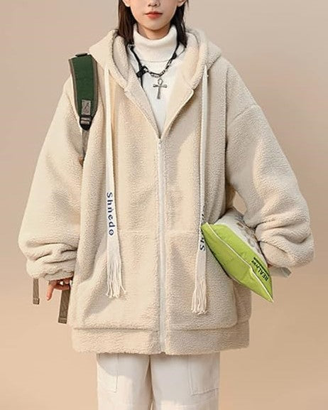 Frau in beiger Teddyfleece-Jacke mit Kapuze, weißem Rollkragenpullover und grünem Rucksack.