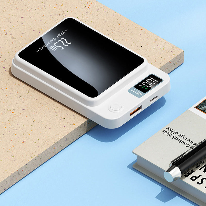 Vit bärbar laddare liggande på en skiva, med USB-A-port, USB-C-port, strömknapp och batteriskärm synliga, bredvid en bok och en penna. 6