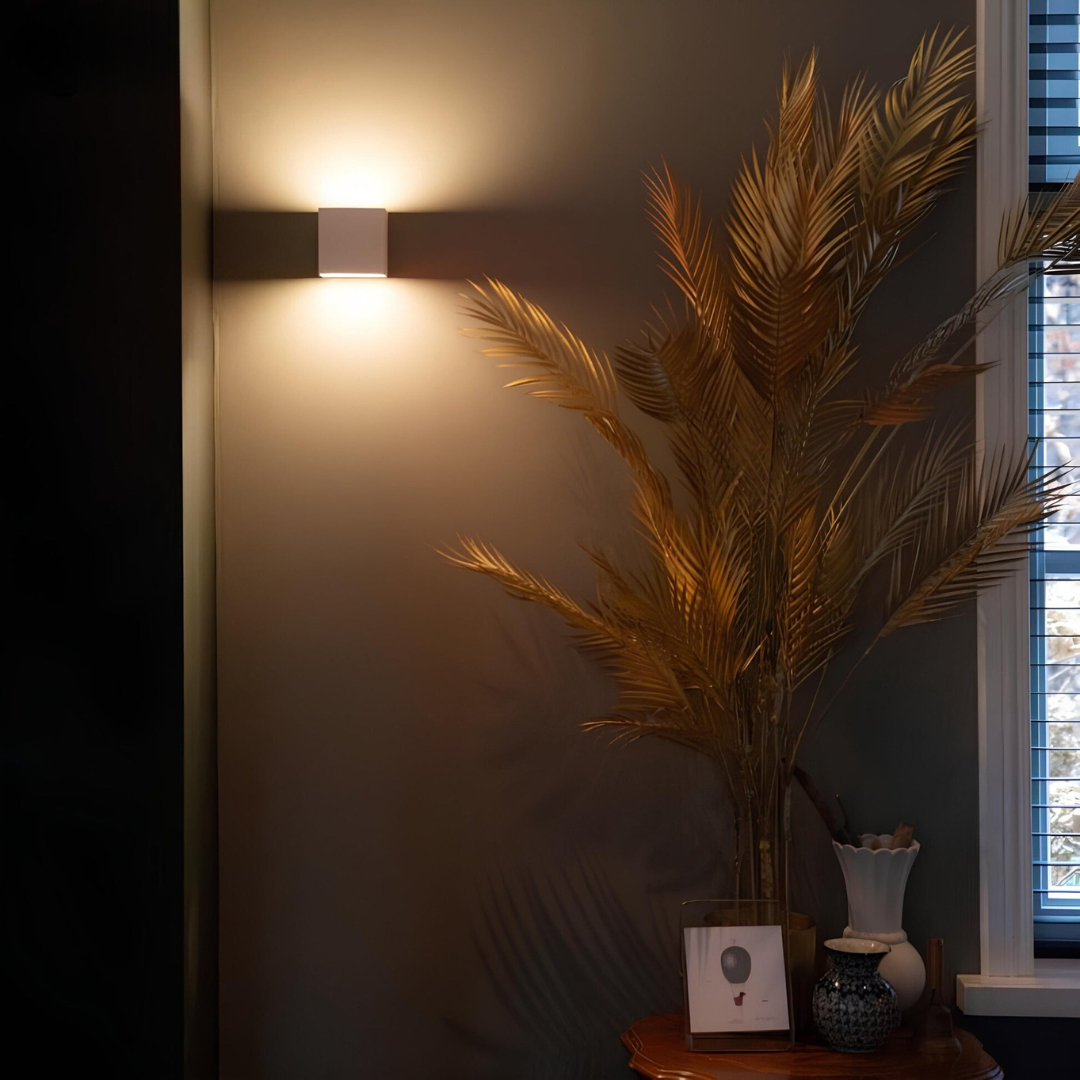 Vit kubformad vägglampa på en mörk vägg bredvid en vas med gyllene palmblad och ett fönster. 4