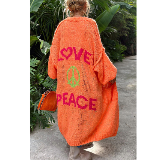 Frau im orangefarbenen Strickmantel mit "Love Peace"-Aufdruck, tropische Pflanzen im Hintergrund.
