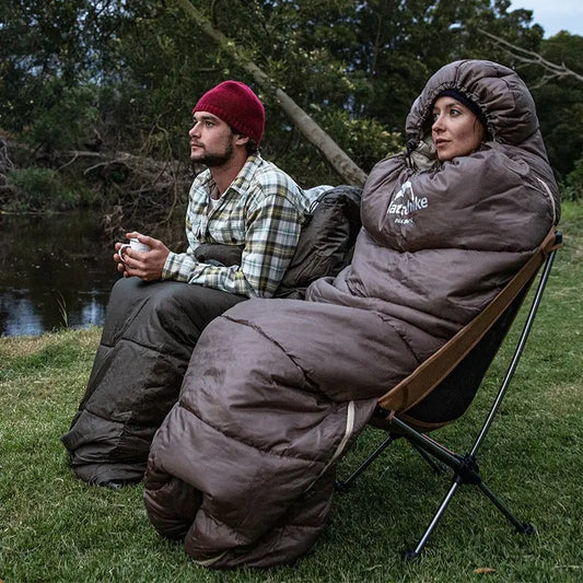 Zwei Personen in Schlafsäcken, Campingstuhl, Outdoor, Natur, Flussufer, Abend, Freizeitkleidung.