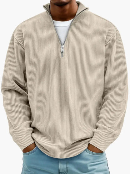 Man i beige ribbstickad tröja med dragkedjekrage, vit t-shirt under och händerna i fickorna på ljusblå jeans. 4