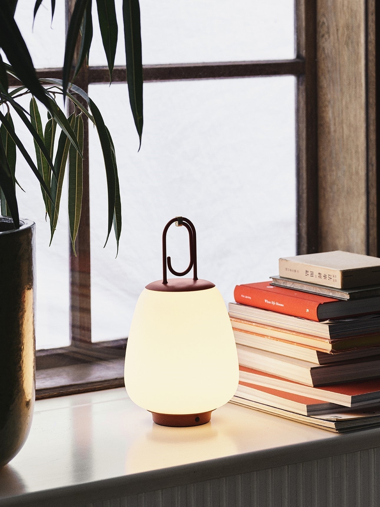 Tragbare LED-Lampe auf Fensterbank, weißer Schirm, roter Griff, neben Büchern, modernes Design.