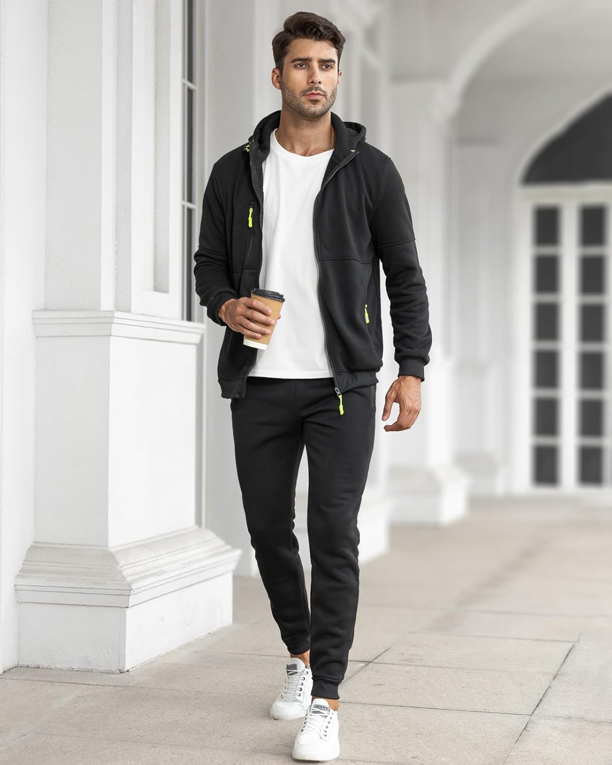 Mann in schwarzem Jogginganzug mit Kapuze, weißem T-Shirt und Sneakers, hält Kaffeebecher.