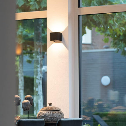 Svart vägglampa på en vit pelare mellan stora fönster, med dekorativa skulpturer och en kruka i förgrunden. 8