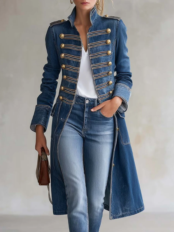 Närbild framifrån av blå denimkappa med guldfärgade knappar och galonband, buren med jeans och vit topp, handen i fickan. 1