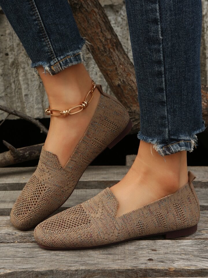 Bruna stickade slip-on-skor med genombrutet mönster sedda från sidan på en modell i mörka jeans, stående på trädäck. 5
