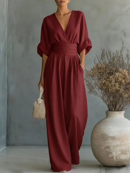 Jumpsuiten i mörkt vinrött med V-ringning och vida ben, buren av en kvinna framför grå vägg. 4
