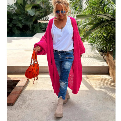 Frau in pinkem Strick-Cardigan, weißem T-Shirt, Jeans und Stiefeln, hält orange Tasche.