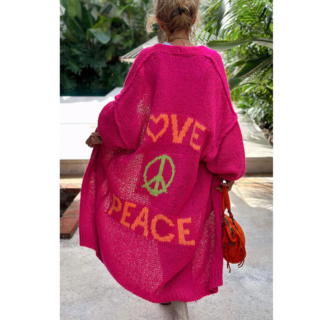Frau in pinkem Strickmantel mit "Love Peace" Schriftzug, tropische Umgebung.
