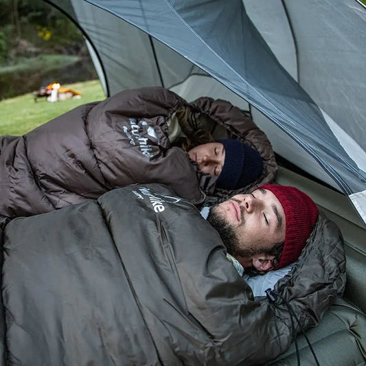 Zwei Personen in braunen Schlafsäcken im Zelt, Camping, Outdoor-Abenteuer, warme Kleidung.