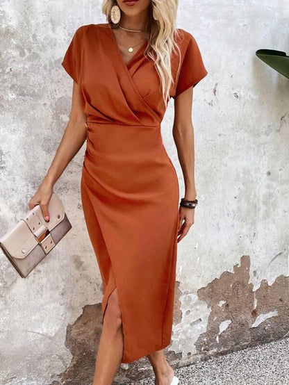 Frau in elegantem, orangefarbenem Wickelkleid mit Clutch vor rustikaler Wand.