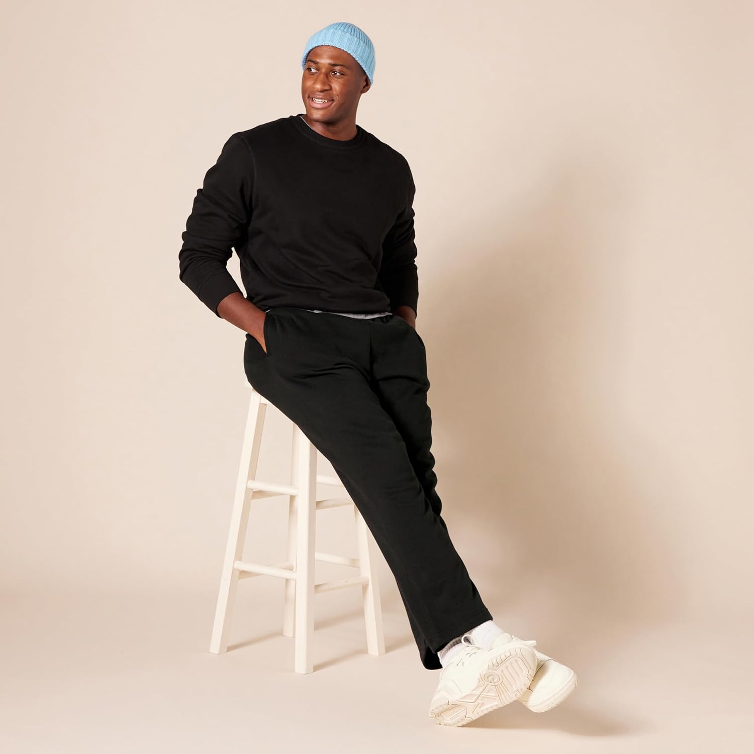 Mann in schwarzem Outfit auf weißem Hocker, blaue Mütze, weiße Sneaker, beige Hintergrund.