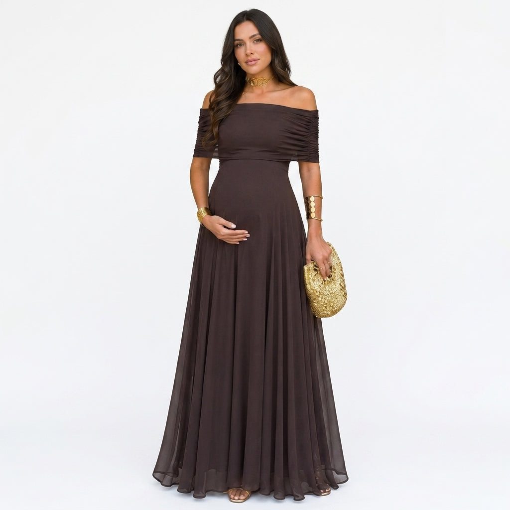 Gravid modell i mörkbrun off-shoulder-maxiklänning med rynkade ärmar, framifrån mot vit bakgrund, med guldväska 1