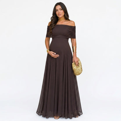 Gravid modell i mörkbrun off-shoulder-maxiklänning med rynkade ärmar, framifrån mot vit bakgrund, med guldväska 1
