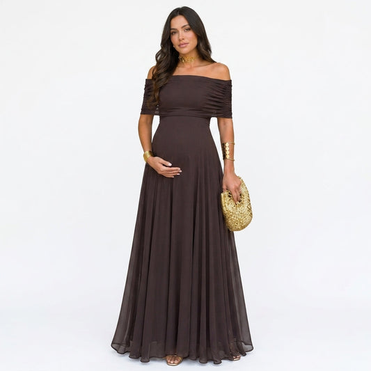 Gravid modell i mörkbrun off-shoulder-maxiklänning med rynkade ärmar, framifrån mot vit bakgrund, med guldväska 1